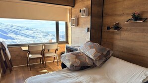 Een slaapkamer met een groot raam, houten wanden, een bed met gekleurde kussens, een bureau en stoelen.
