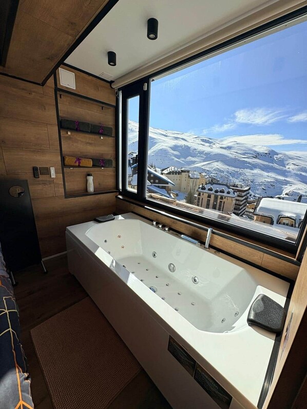 A modern bathroom with a large bathtub, wooden wall paneling, a window with a snowy mountain view, and a shelf with toiletries.