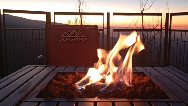 A barbecue grill with flames, a branded bag, and a view of the sunset over a fence. - SnowGranada Los Valles Alodia Parking+Chair (Sierra Nevada)