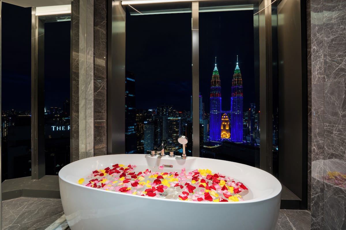 Une salle de bain moderne avec une grande baignoire remplie de pétales de roses, un mur en marbre et une vue sur un paysage urbain avec des bâtiments illuminés la nuit.