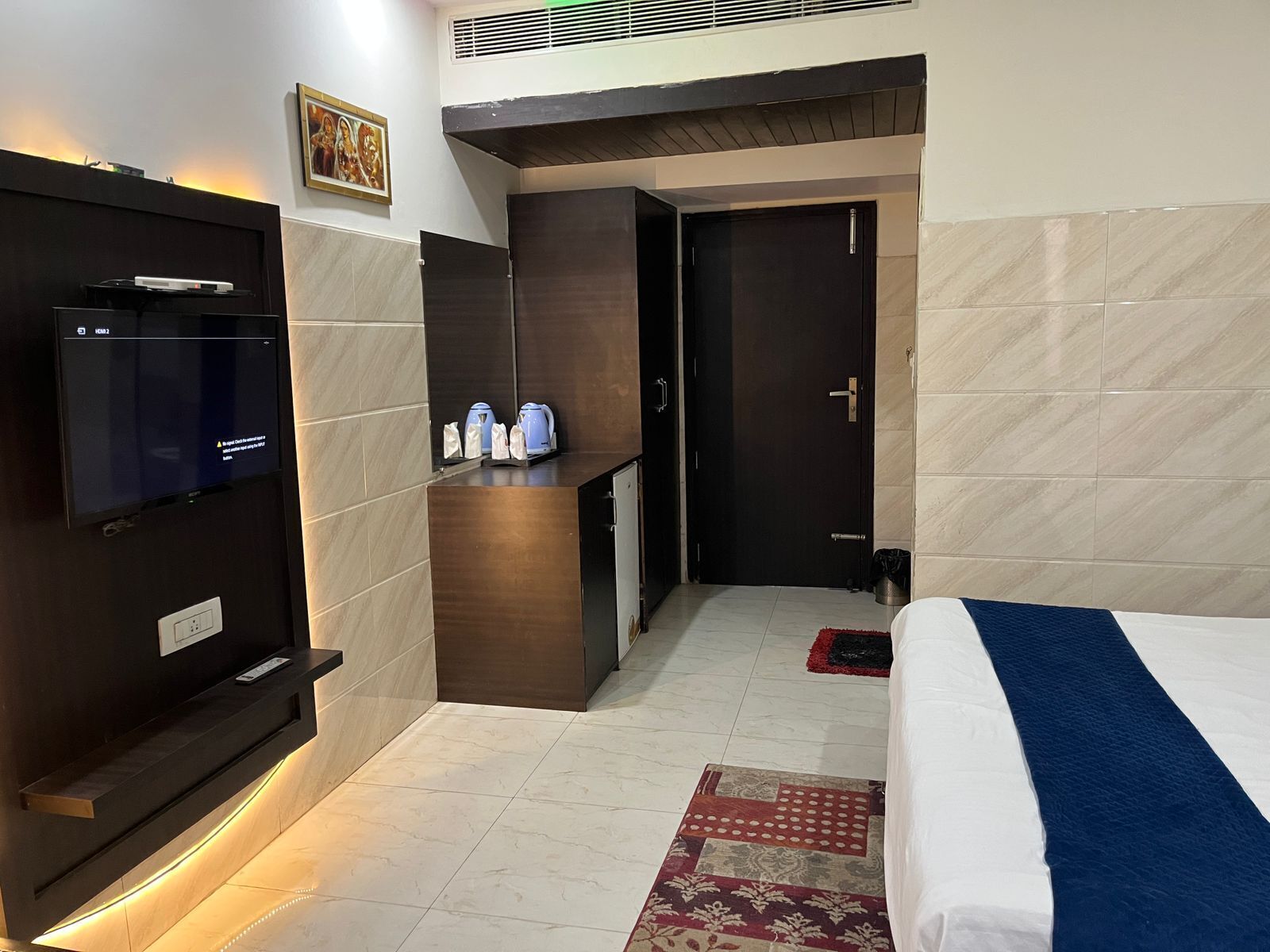 A hotel room with a bed, a TV mounted on the wall, a small desk with a kettle, and a door leading to another room.