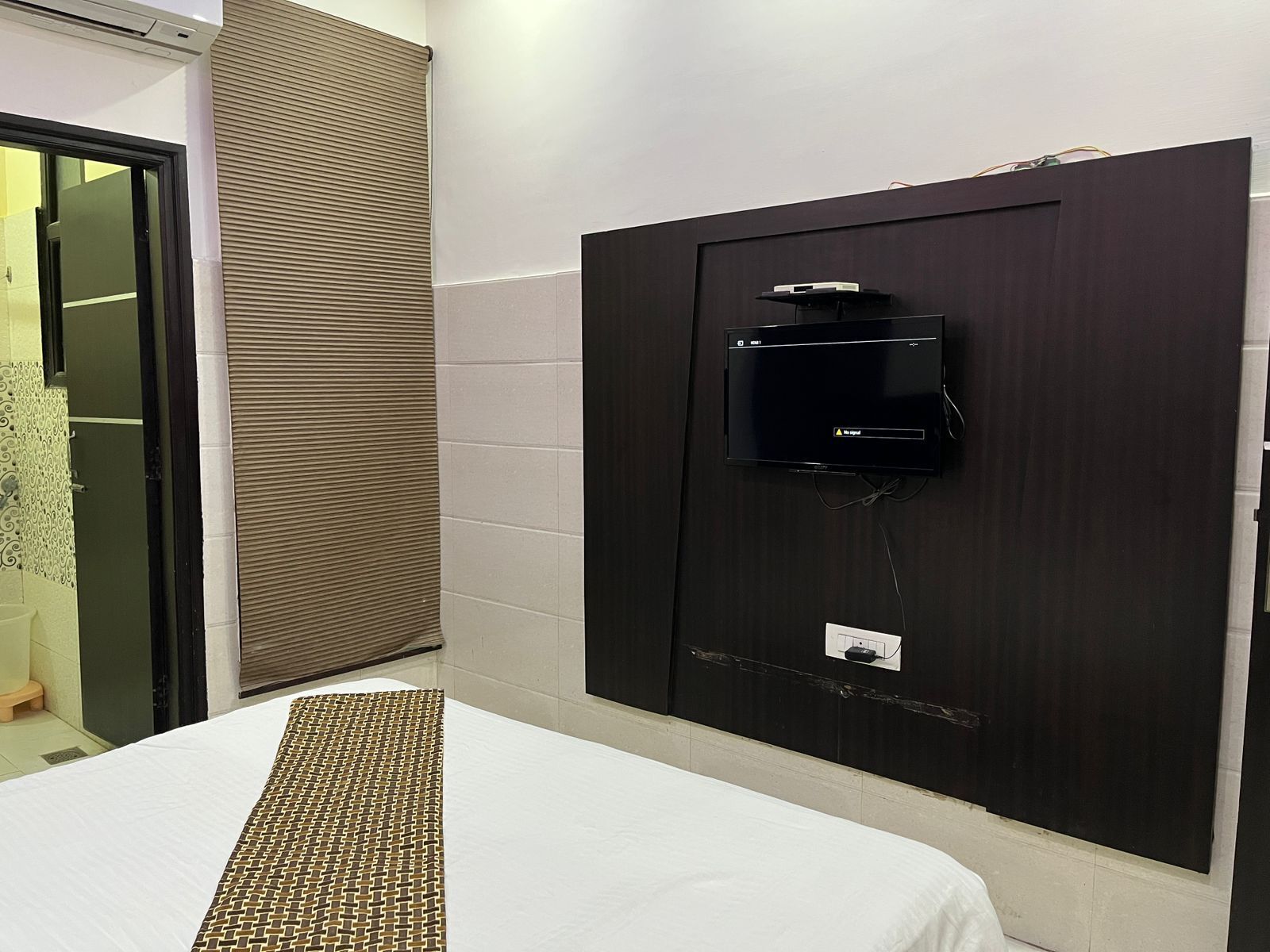 Habitación de hotel con cama, televisor montado en una pared oscura, ventana con persianas y una puerta a la izquierda.