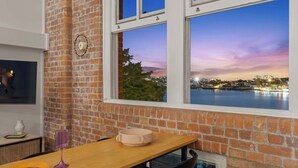 A room with a brick wall, a wooden table, a window with a view of a cityscape at dusk, and a clock on the wall. - Luxury Water Views ~ 2Bed/2Bath/1Car/Pool (Teneriffe)