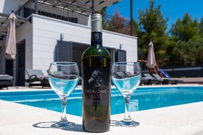 Eine Flasche Frenchie-Wein und zwei Gläser auf einem Tisch am Pool, mit Pool und Gebäude im Hintergrund.