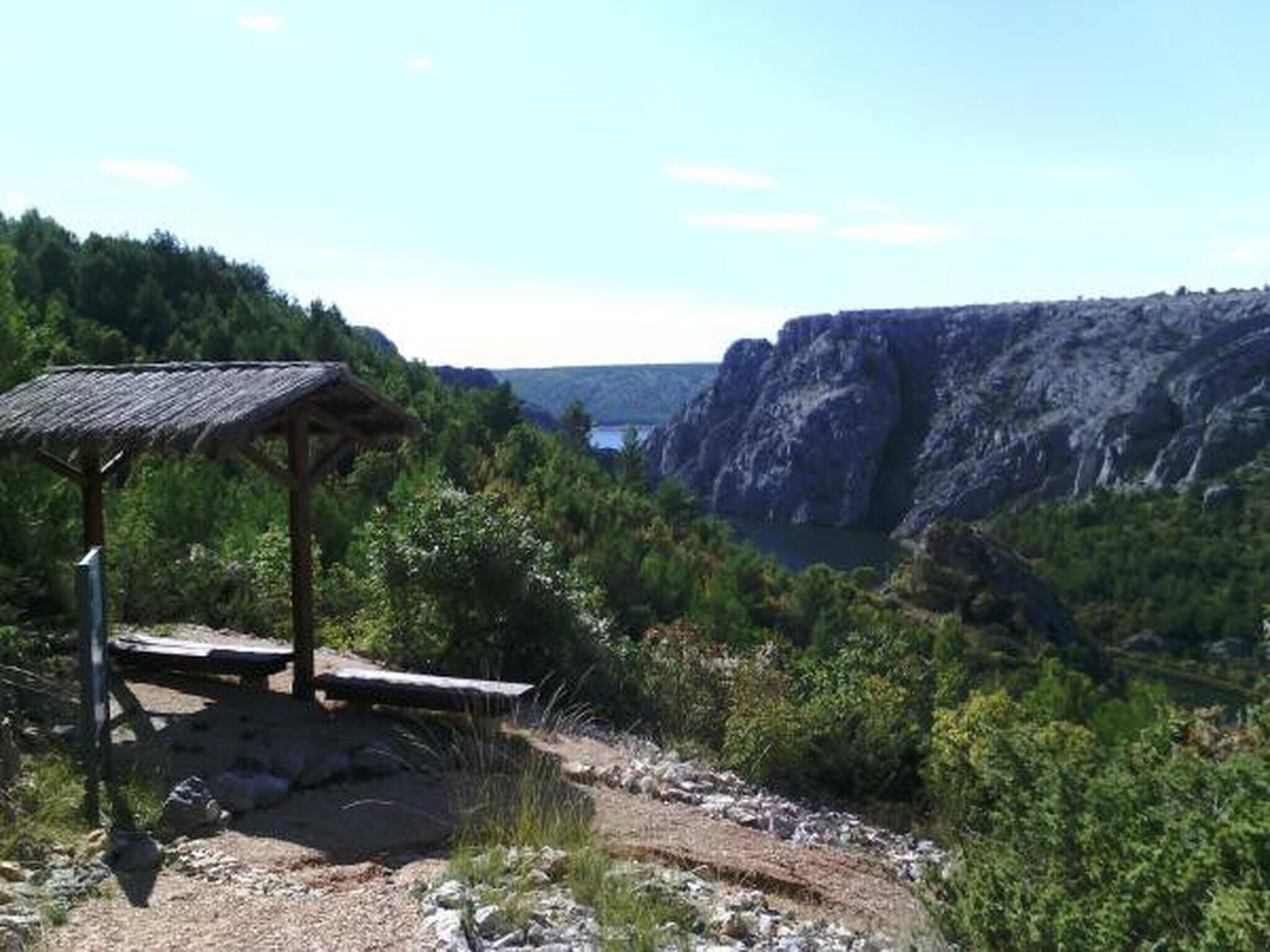 A rustic wooden shelter with a sloped roof, surrounded by rocky terrain and dense greenery, overlooking a steep cliff and a body of water.