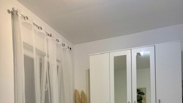 Un dormitorio con cama, un armario blanco con puertas espejadas y una cortina ligera sobre la ventana.