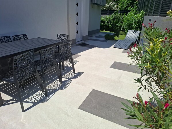 An outdoor patio with a table and chairs, a small garden area, and a pathway leading to a door.