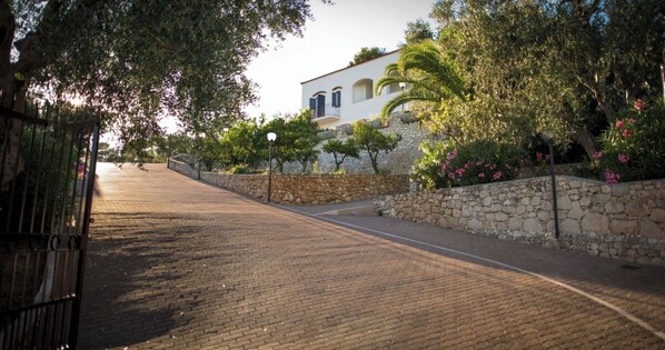 Un chemin pavé mène à un bâtiment blanc aux murs de pierre, entouré d’une végétation luxuriante.