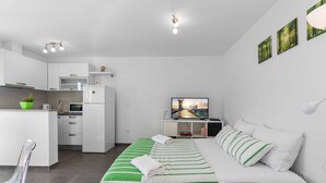 Una habitación moderna y funcional con kitchenette, una cama con ropa de cama a rayas verdes, televisor y un pequeño rincón de estar con una silla.