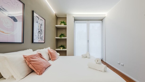 A modern bedroom with a large bed, two wall-mounted artworks, a small shelving unit with plants, and a large window with a view.
