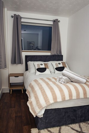 A bedroom with a bed, a nightstand, a window with curtains, and a wooden floor.