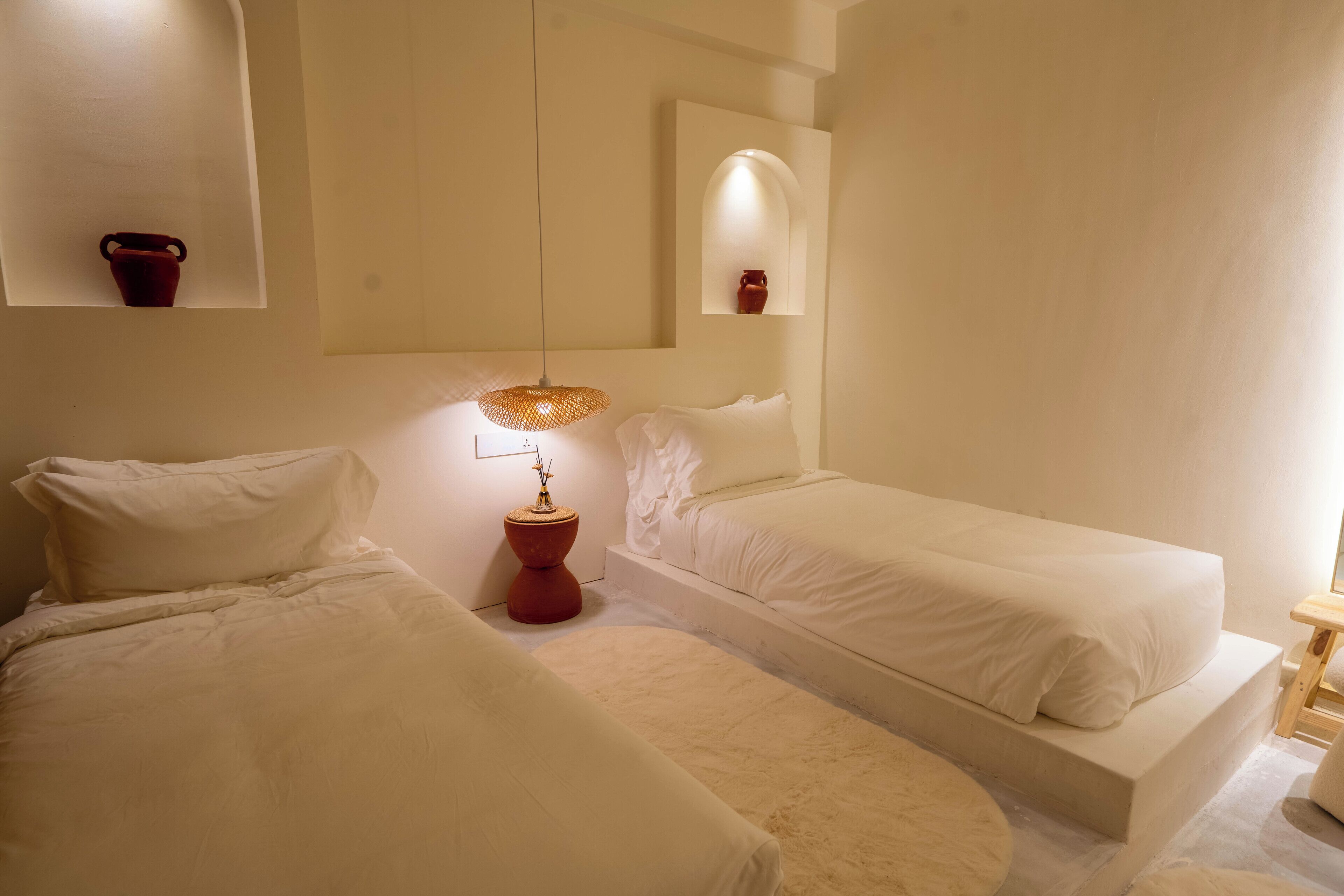 A bedroom with two single beds, a bedside table with a lamp, a small mirror, and a red vase on the wall.