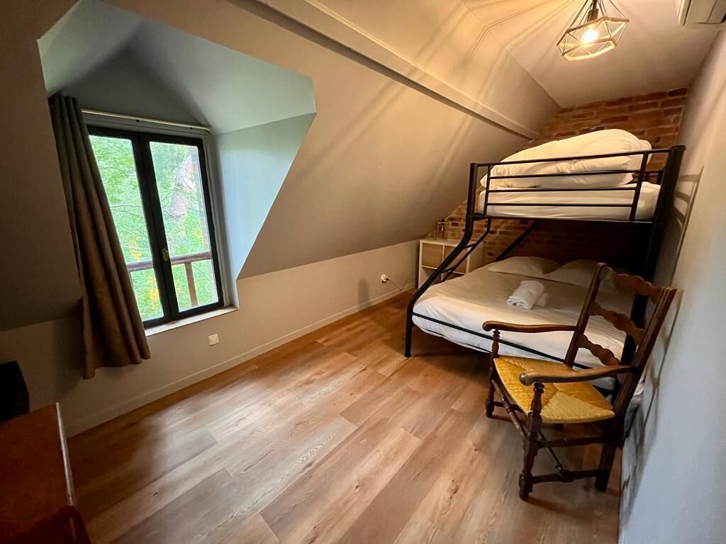A dormitory room with a bunk bed, a wooden chair, a window with curtains, and a hanging light fixture.