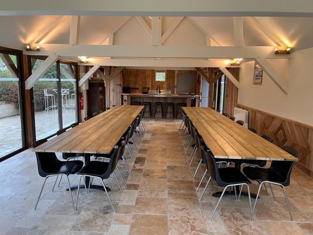 A large wooden table with black chairs in a spacious room with stone flooring and large windows.