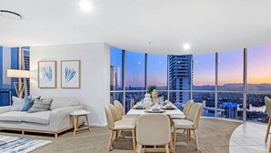 Una sala de estar moderna con comedor, ventanas amplias y vistas a la ciudad, y un sofá blanco con cojines azules.