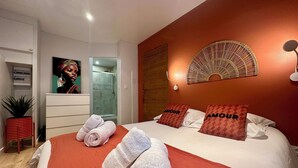 Ein Hotelzimmer mit einem Bett, zwei Kissen mit dem Aufdruck 'AMOUR', einem Wandventilator, einer Dusche und einem Bild an der Wand.