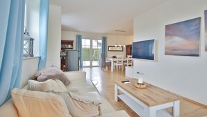 Ein modernes Wohnzimmer mit einer Couch, einem Holztisch und einem Fernseher. Es gibt einen Essbereich mit Tisch und Stühlen sowie eine Küche im Hintergrund.