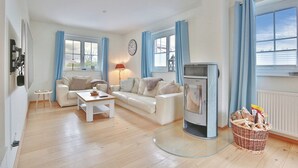 Ein Wohnzimmer mit einem weißen Sofa, einem Couchtisch, einem Fernseher und einem Kamin. Zwei Fenster mit blauen Vorhängen und ein Korbkorb mit Holzscheiten in der Nähe des Kamins.