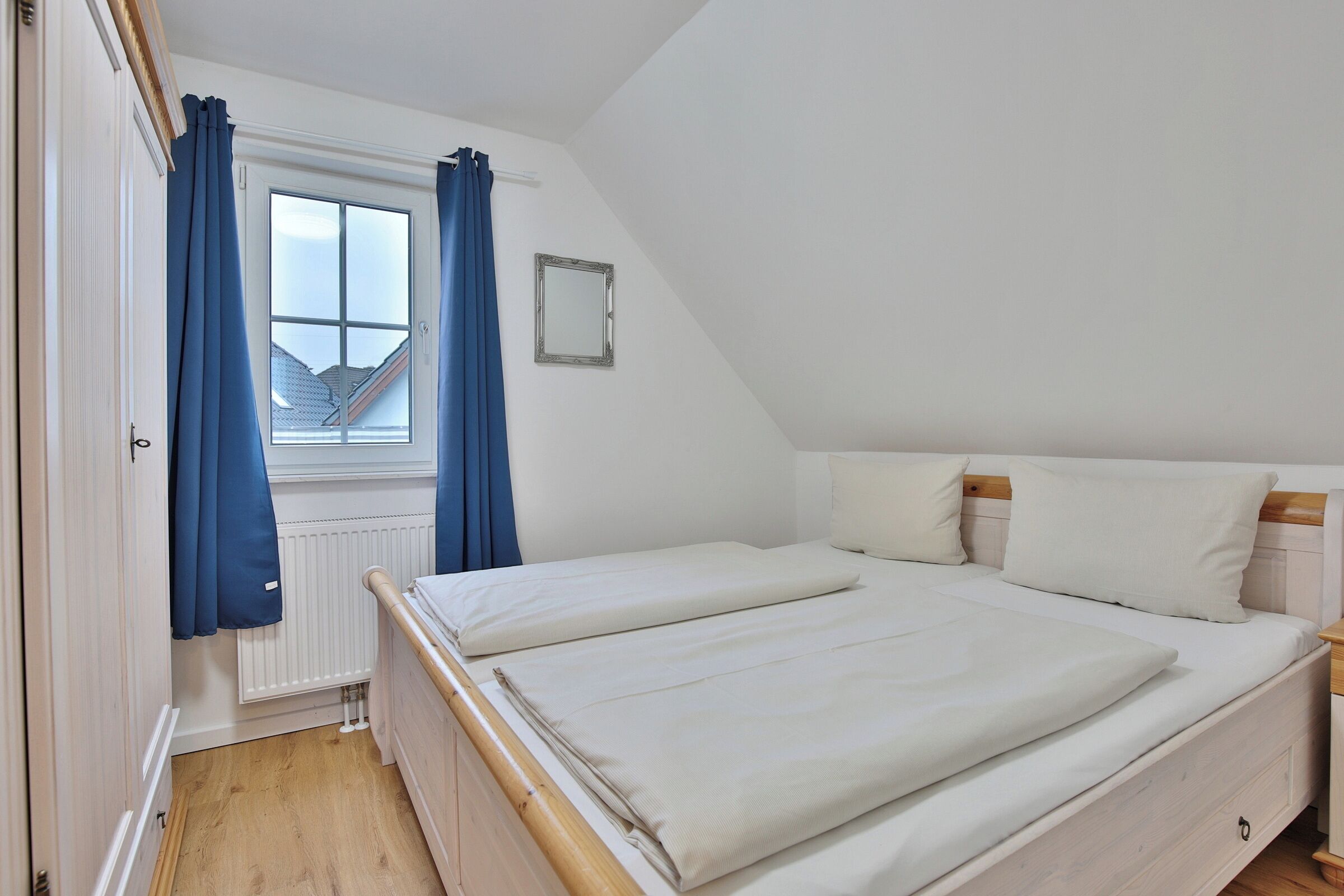 A bedroom with a bed, a window with curtains, a radiator, and a small mirror on the wall.