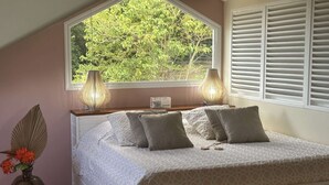 Ett sovrum med ett stort fönster, en säng med mönstrat sänglinne, två sänglampor och en vas med blommor.