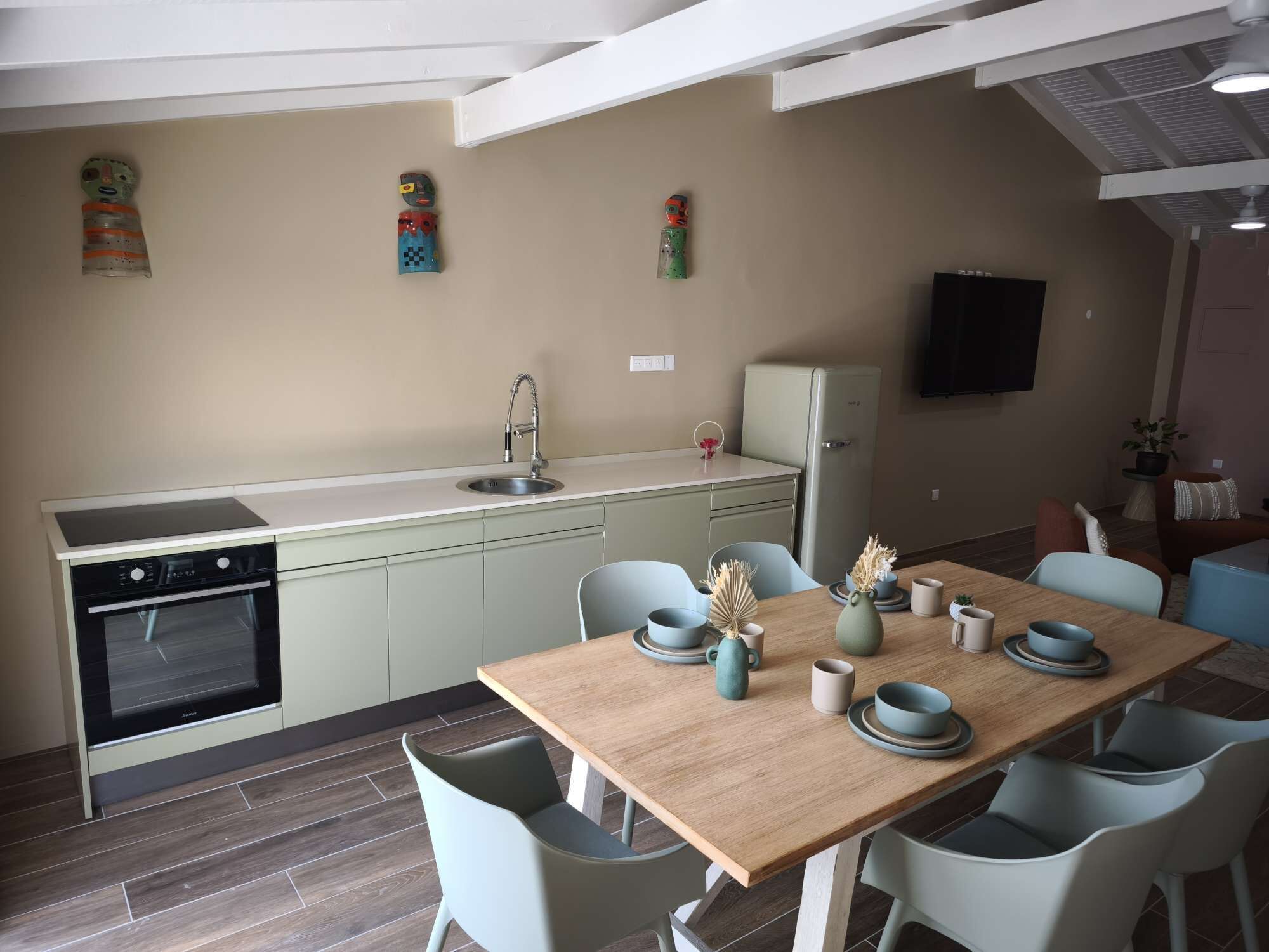 Cozinha compacta com forno embutido, pia e geladeira. Há uma mesa de jantar com cadeiras e uma TV fixada na parede.