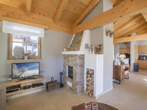 A cozy living room with a stone fireplace, wooden beams, a television, and a kitchen in the background.