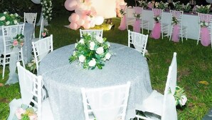 Een bruiloftdecoratie buiten met witte stoelen en tafels, een bloemenboog en een witte gebouw op de achtergrond in de schemering.