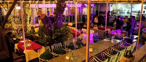 An outdoor dining area with tables and chairs, surrounded by string lights and a pergola structure.