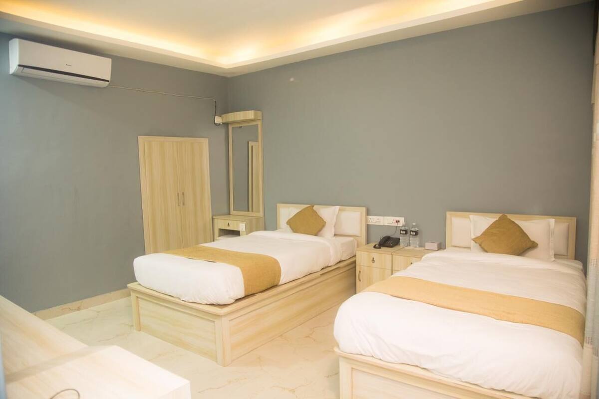 A hotel room with two beds, a wardrobe, a bedside table with a phone and toiletries, and an air conditioning unit.