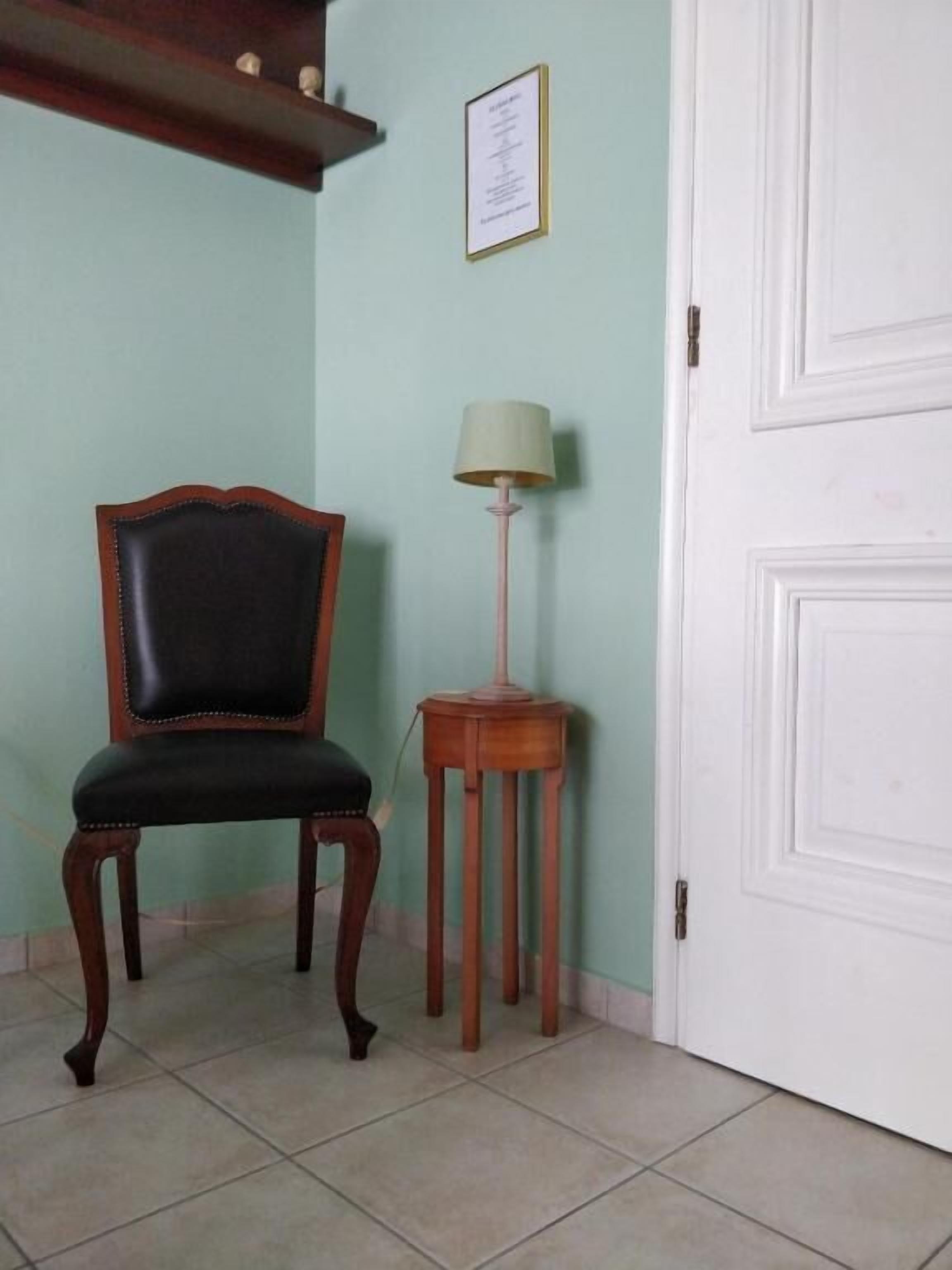 Una habitación con una silla de madera, una pequeña mesa de madera con una lámpara y una puerta blanca.