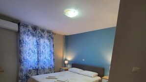 A neatly made bed with a white comforter and pillows, a bedside lamp, and a window with blue curtains.