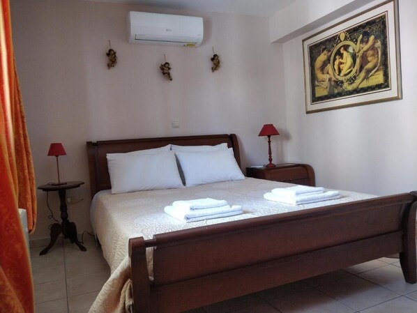 A bedroom with a wooden bed, white linens, two bedside tables with lamps, an air conditioner, and a framed painting on the wall. - Sior Dionysios Apartments (Kefalonia)