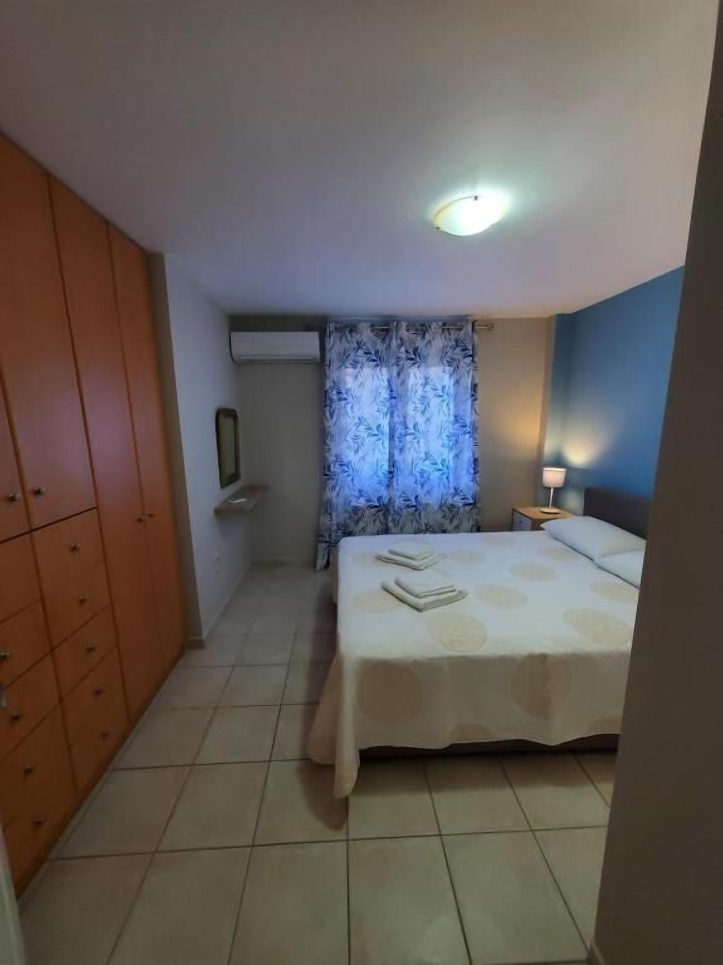 A bedroom with a bed, a wardrobe, a window with curtains, a bedside table with a lamp, and an air conditioning unit.