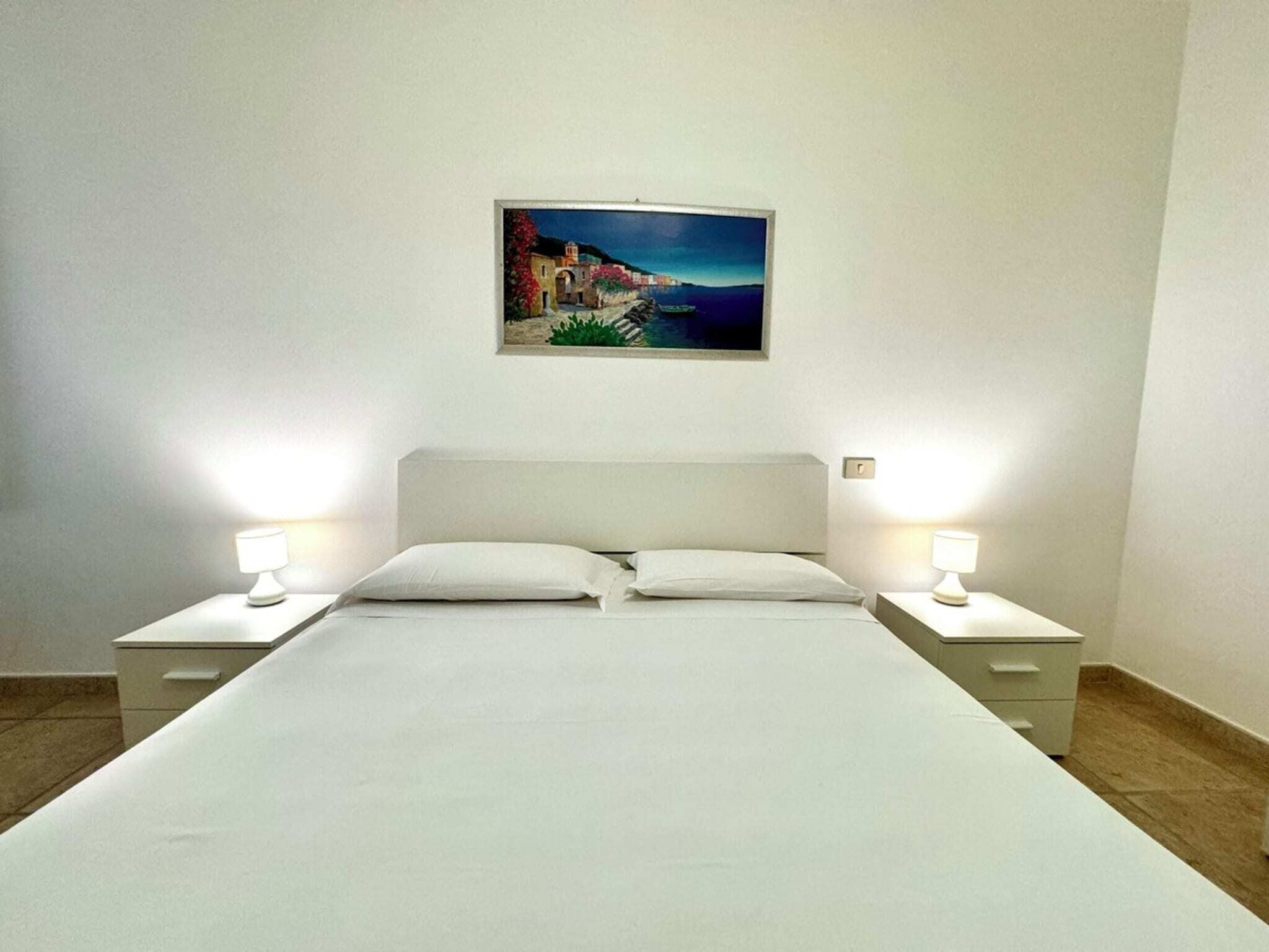 A neatly made bed with white linens and two bedside tables with lamps, a painting of a coastal scene on the wall, and a simple, clean room layout.
