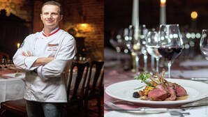 A chef in a white uniform with a badge, standing in a restaurant with a brick wall background.