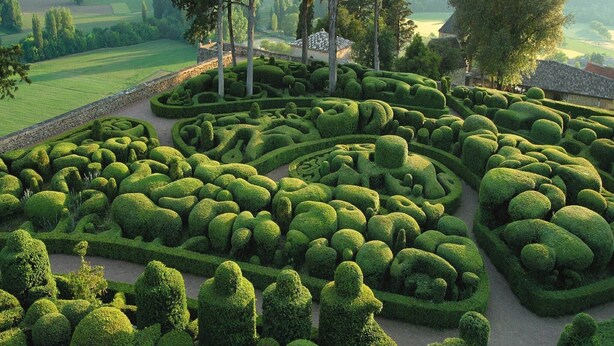 A meticulously landscaped garden with geometric hedges and a rustic building in the background.