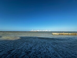 Vista costera con un espigón de piedra y el horizonte de una ciudad a lo lejos.