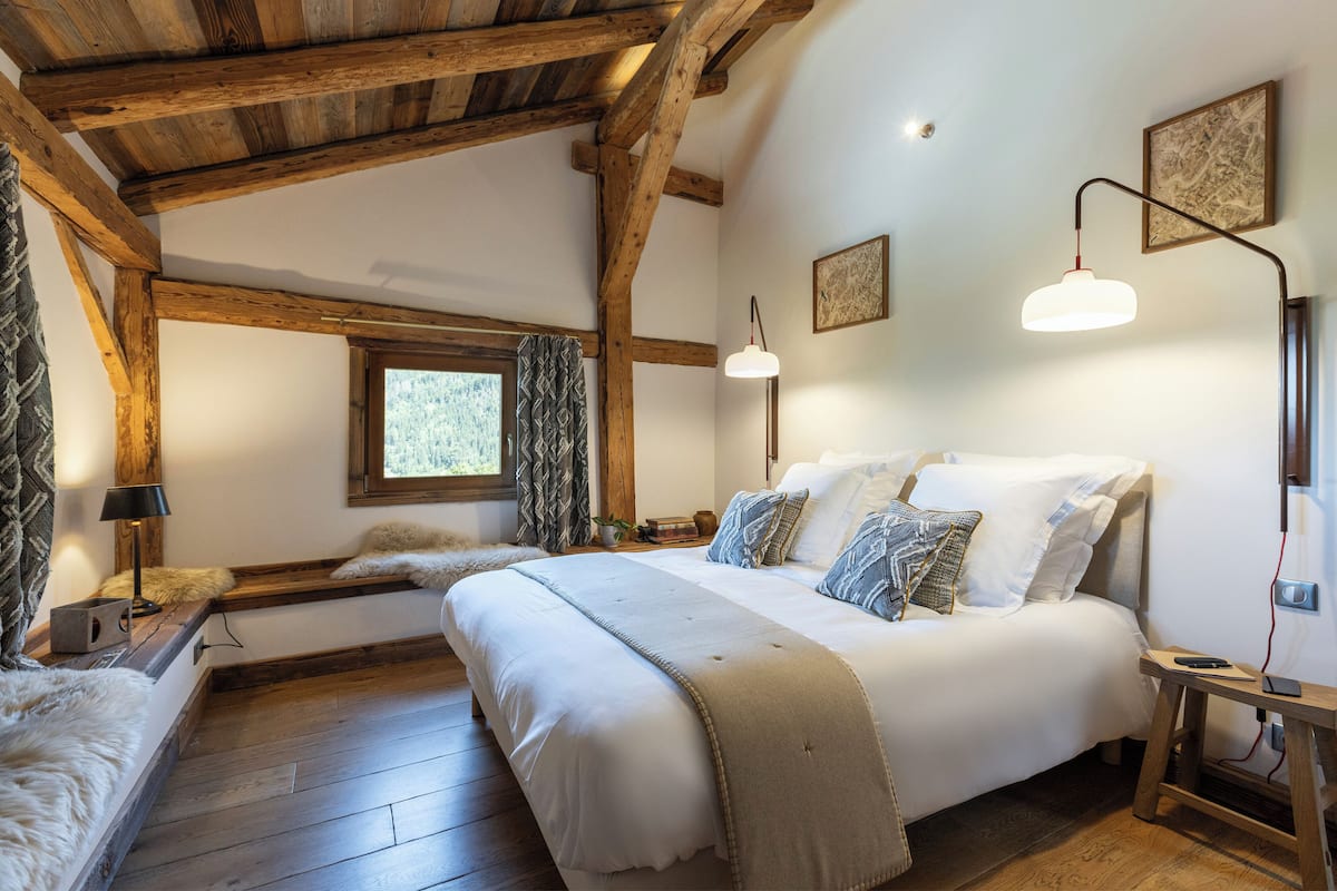 Ein Schlafzimmer mit einem großen Bett, Holzbalken, einem Fenster mit Vorhängen und zwei Wandleuchten.