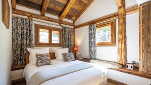 A bedroom with a large bed, wooden beams, patterned curtains, and a window with a view of a house.