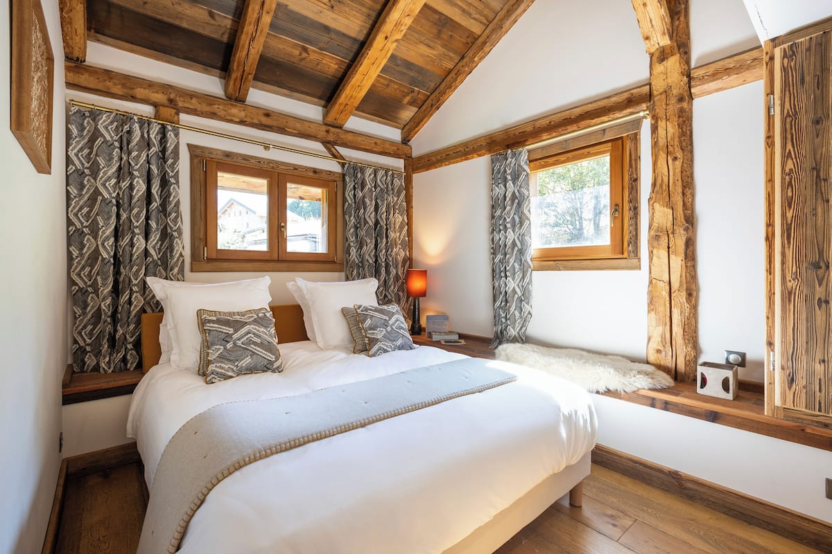 Ein Schlafzimmer mit einem großen Bett, Holzbalken, gemusterten Vorhängen und einem Fenster mit Blick auf ein Haus.