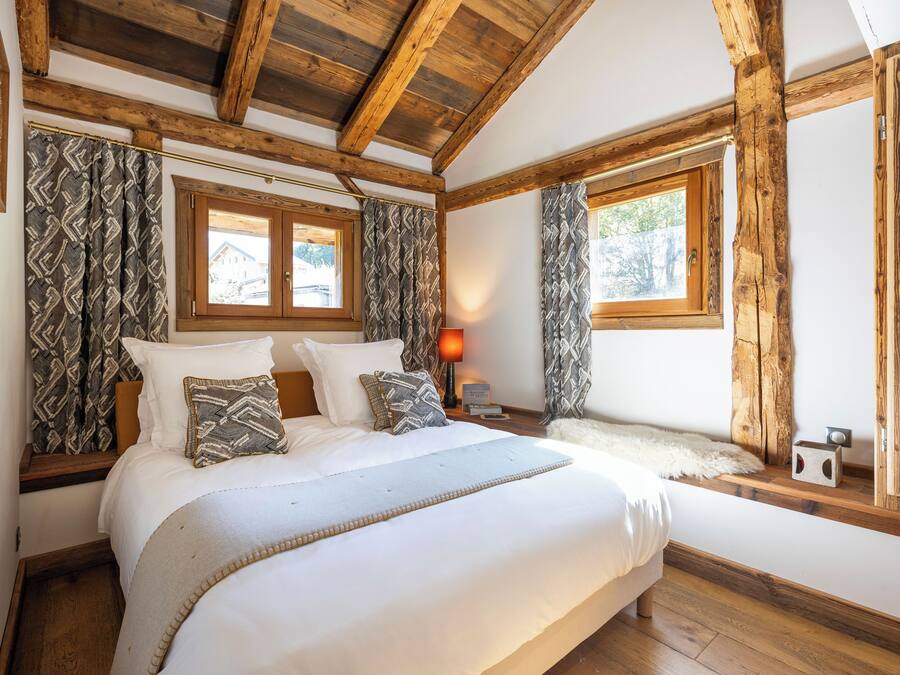A bedroom with a large bed, wooden beams, patterned curtains, and a window with a view of a house.