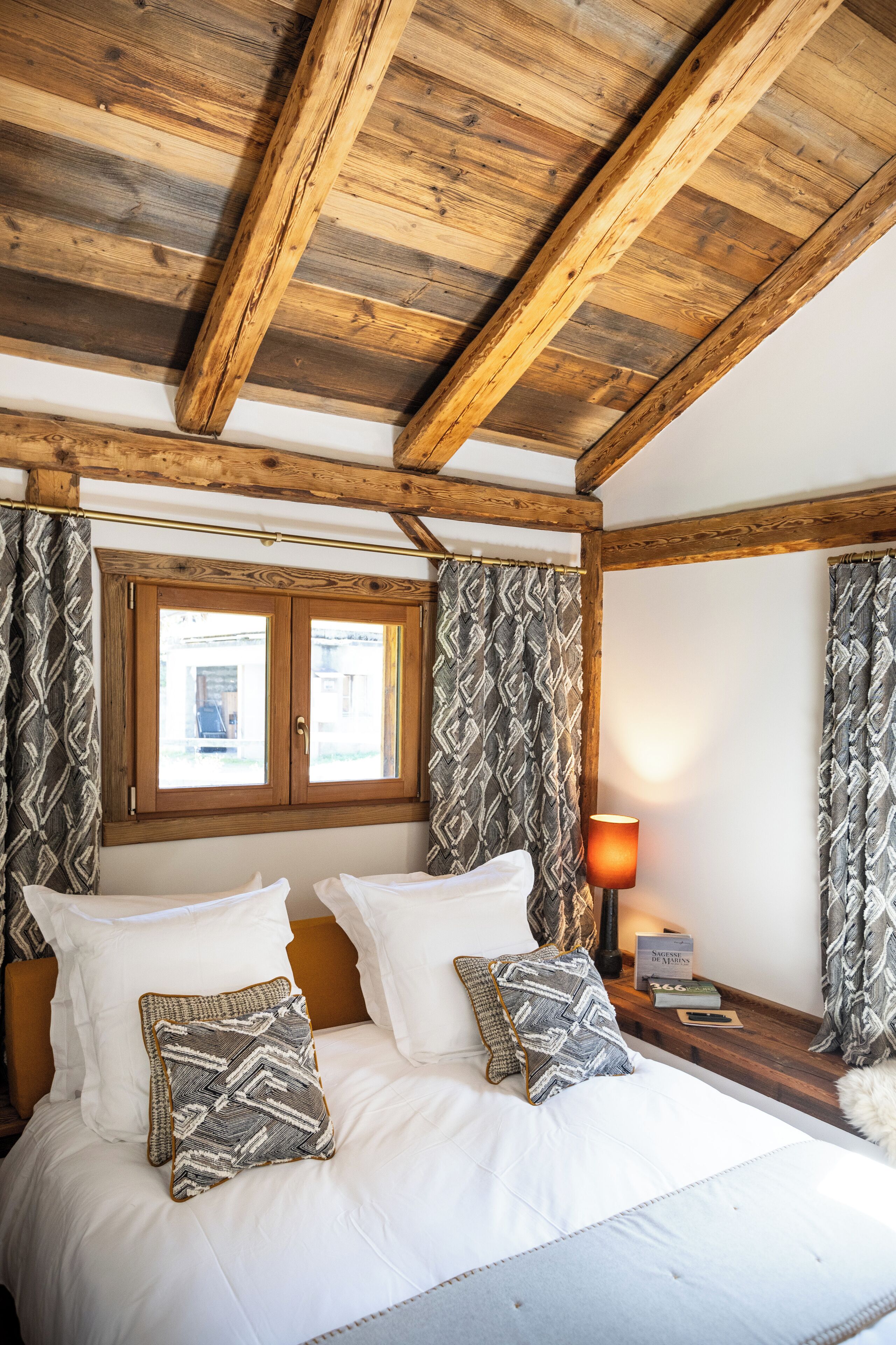 A bedroom with a wooden ceiling, a bed with white bedding, patterned pillows, and curtains with a geometric design.
