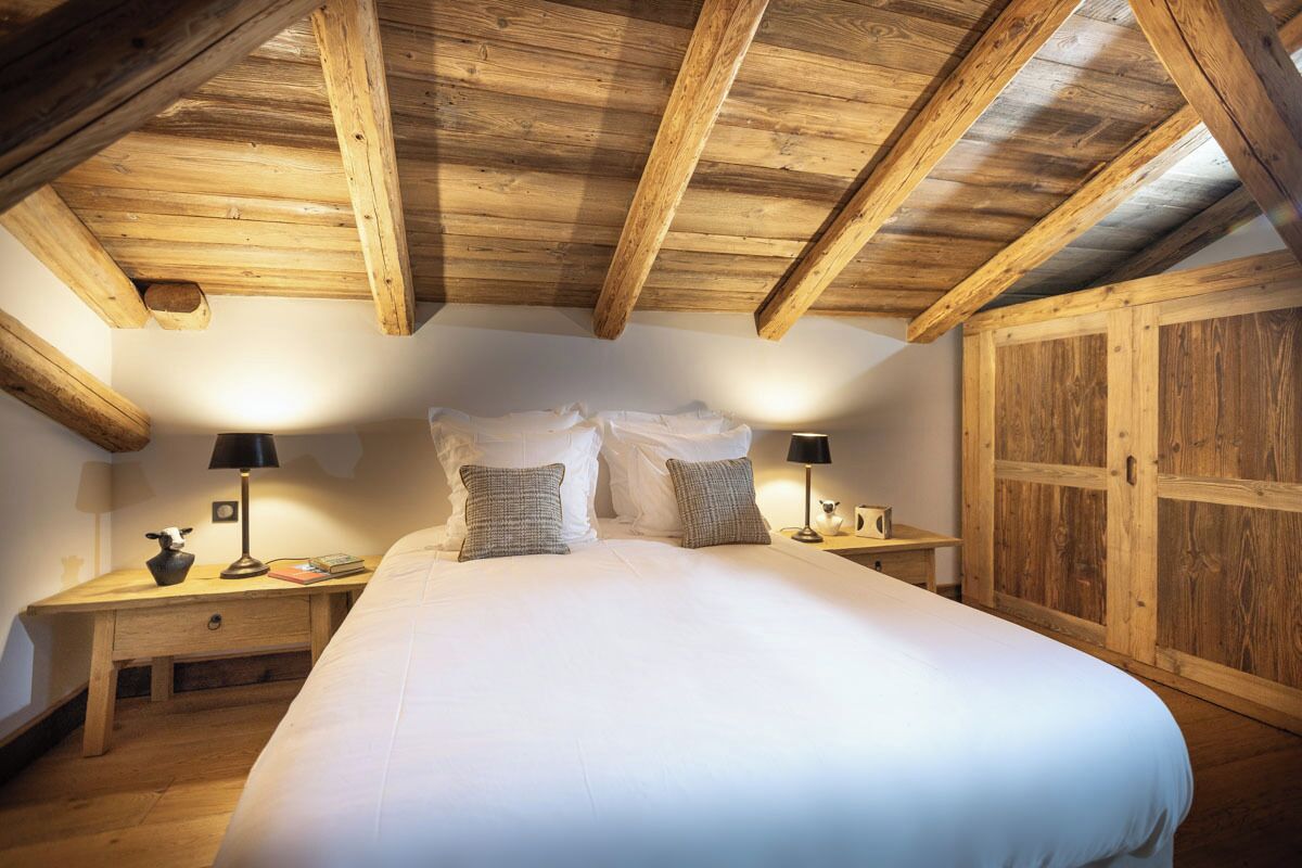 A bedroom with a wooden ceiling, a large bed with white bedding, two bedside tables with lamps, and a wooden wardrobe.