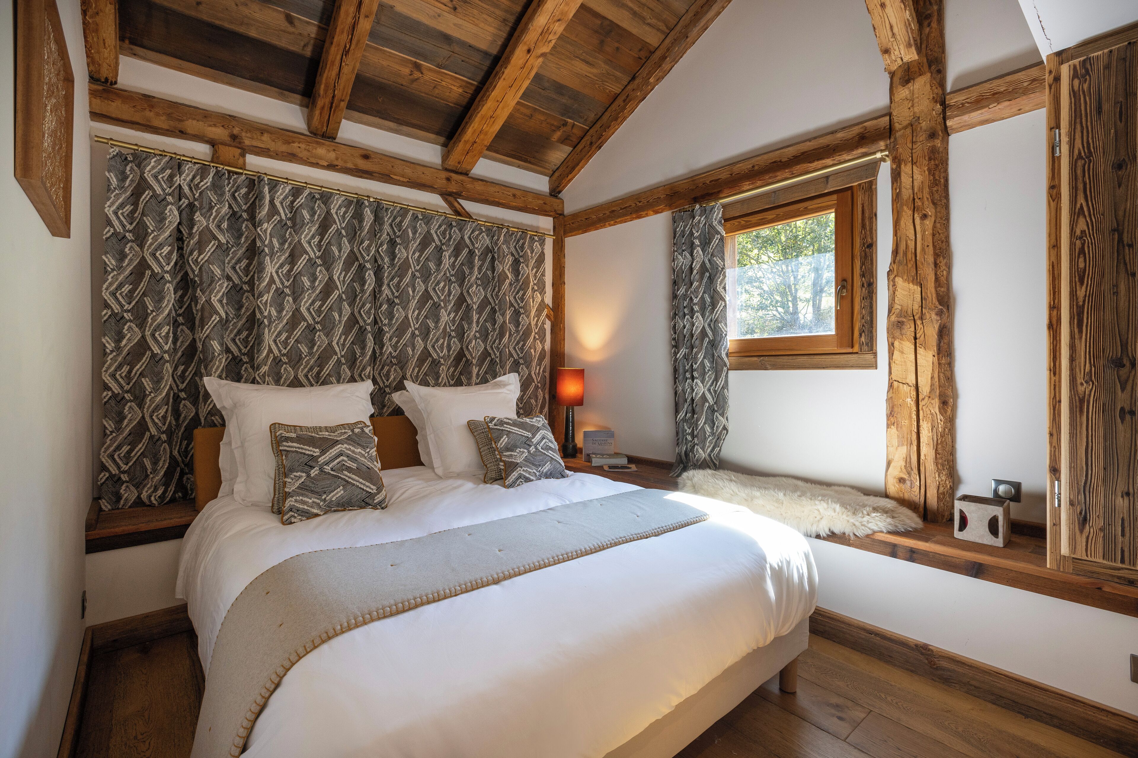 Ein Schlafzimmer mit einem großen Bett, Holzbalken, gemusterten Vorhängen, einem Fenster und einem kleinen Tisch mit einer Lampe.