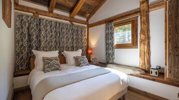 A bedroom with a large bed, wooden beams, patterned curtains, a window, and a small table with a lamp.