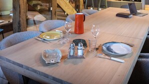 A dining area with a long table set for four, wooden beams overhead, and a kitchen in the background.