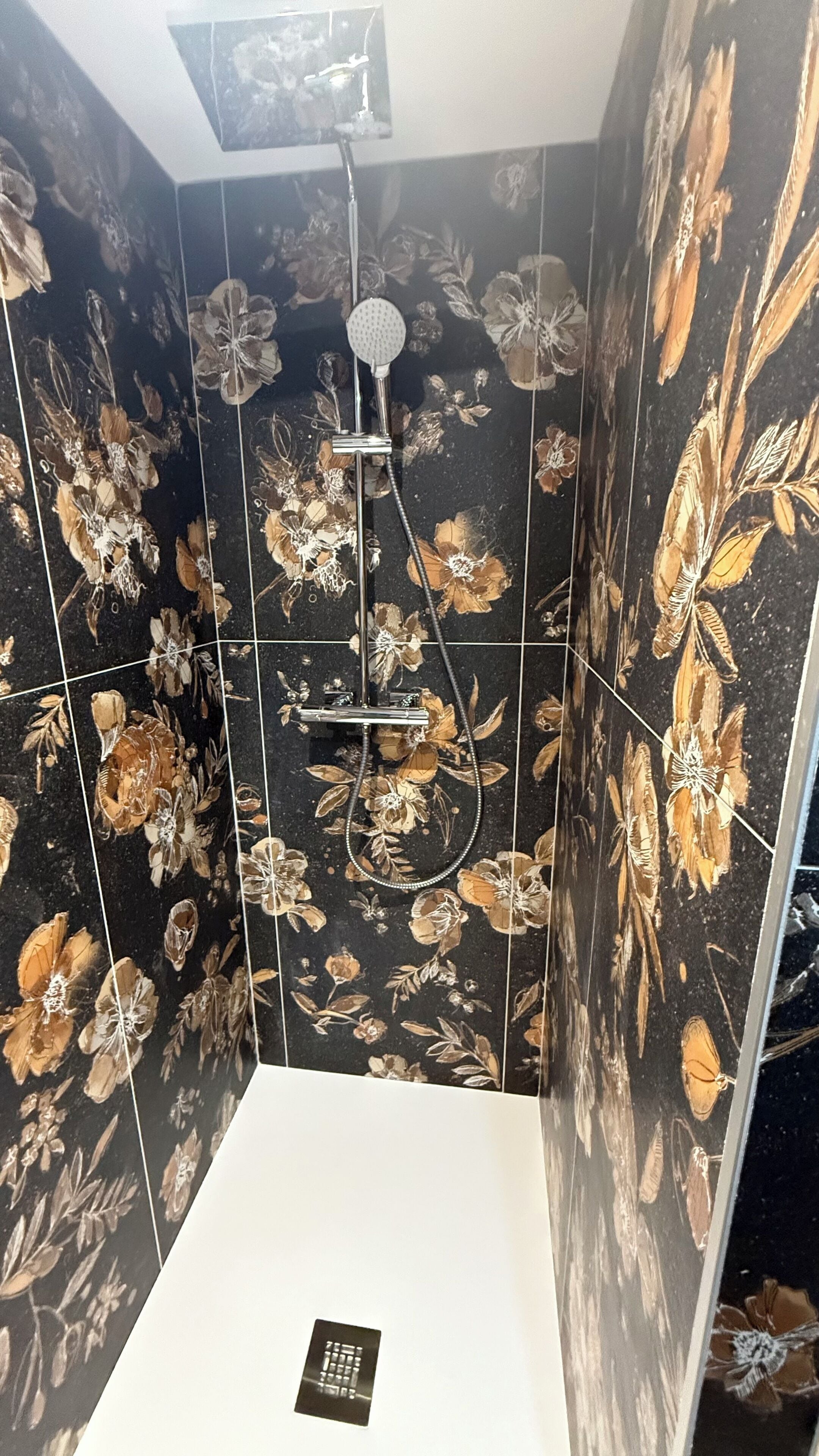 A bathroom with black floral tiles, a shower with a handheld showerhead, and a white sink.