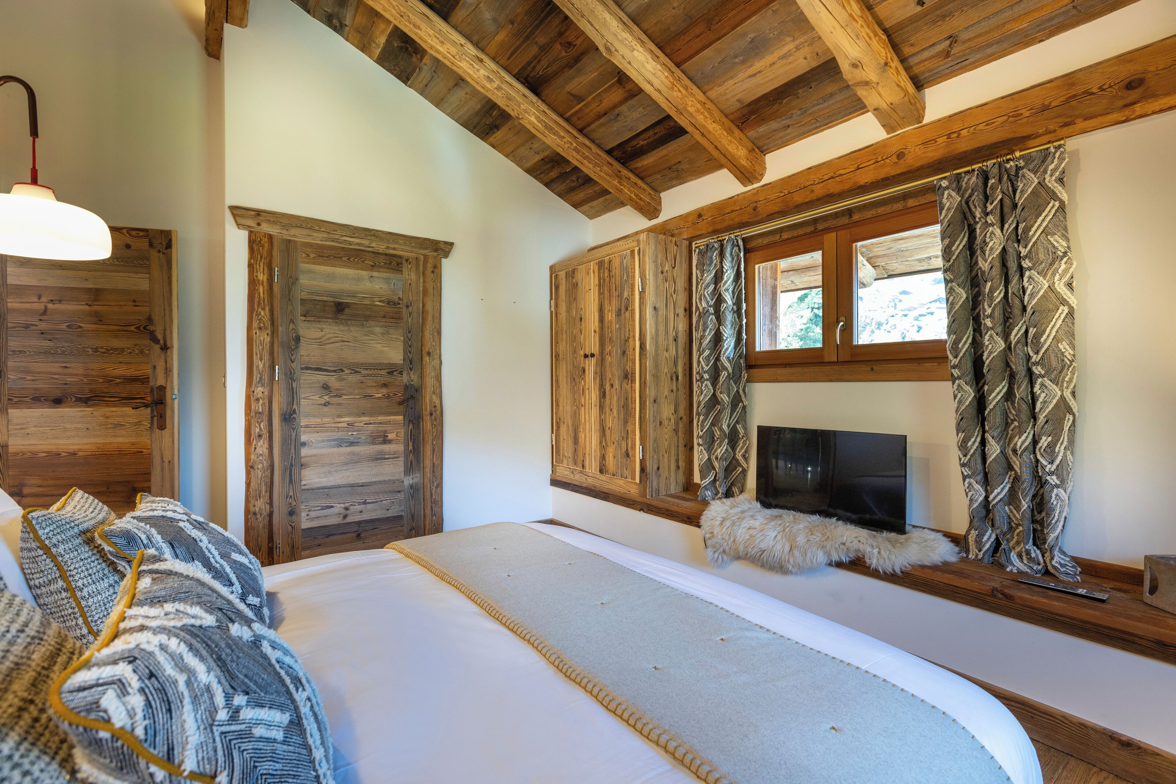 A bedroom with a large bed, wooden ceiling beams, a television, and patterned curtains.