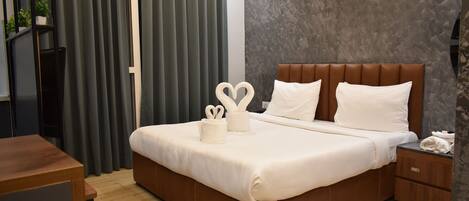 Ein modernes Hotelzimmer mit einem großen Bett, einem Nachttisch aus Holz, einem Schreibtisch und einem dunklen Vorhang.