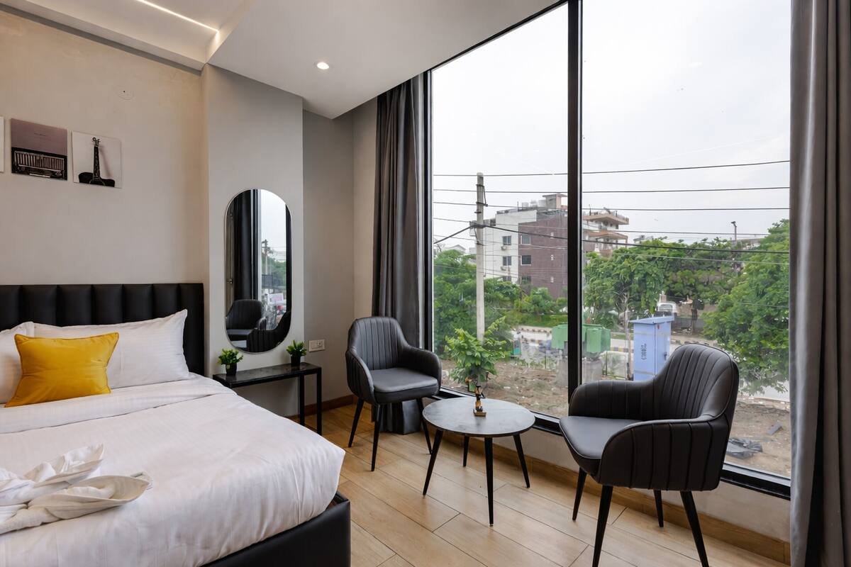 A modern hotel room with a large bed, two armchairs, a small table, and a large window offering a city view.